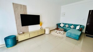 A modern living room with a flat-screen TV, a teal sofa, a small round coffee table, and a beige console with drawers. -  Cozy 3BR Condo with AC at Negombo Santorini Resort, Close to Airport (Negombo)