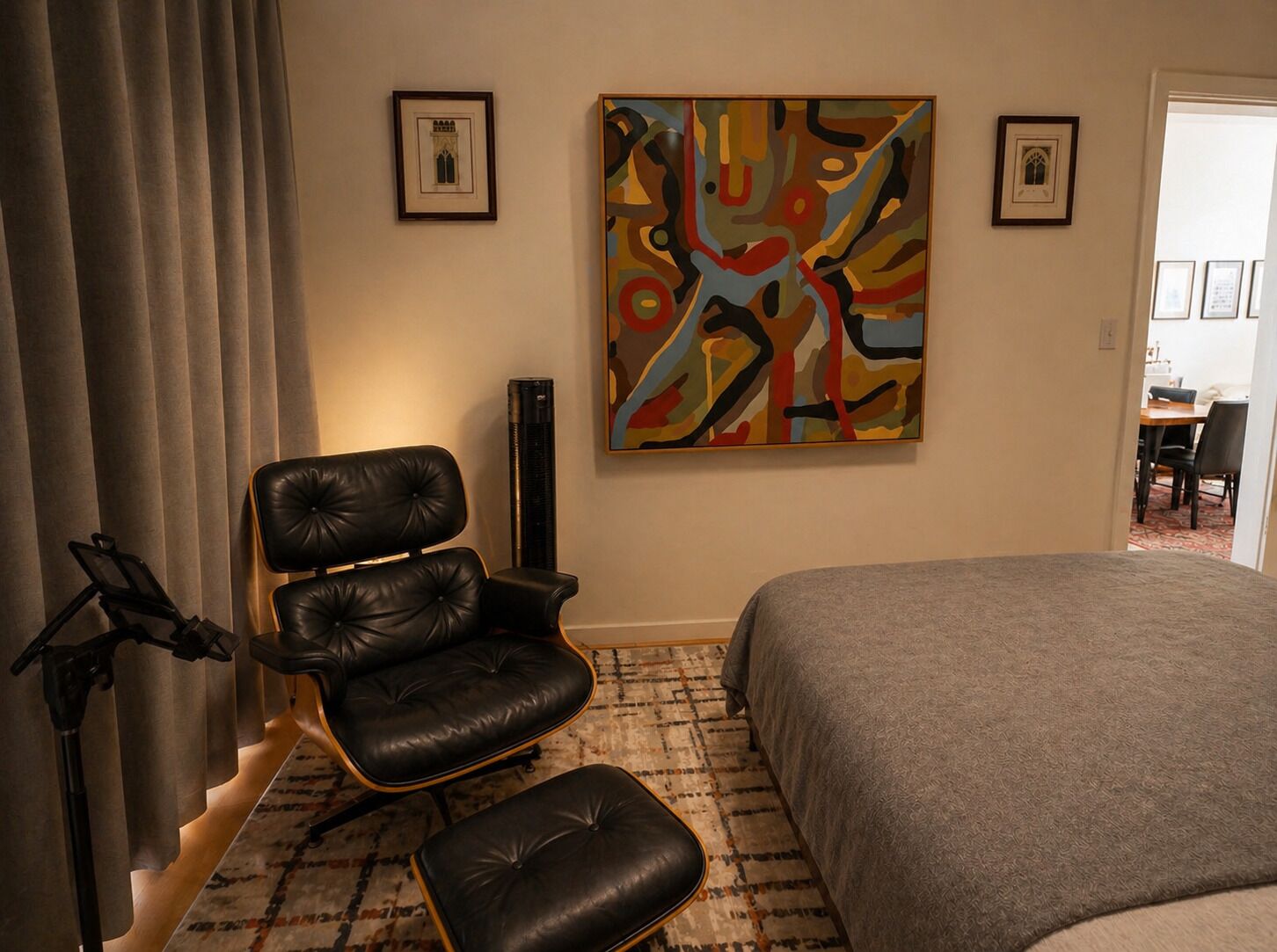 A hotel room with a bed, a black leather chair, a standing lamp, and a colorful abstract painting on the wall.