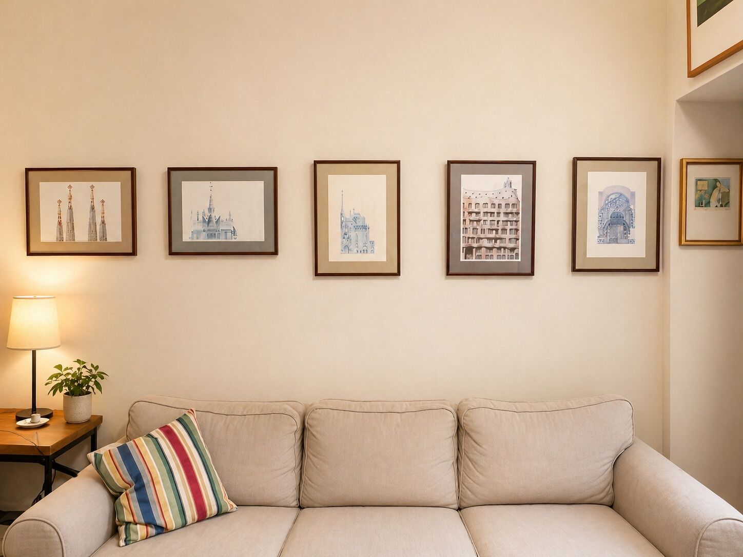 A beige sofa with a striped pillow, a framed picture of a building, a lamp, and a small table with a plant.