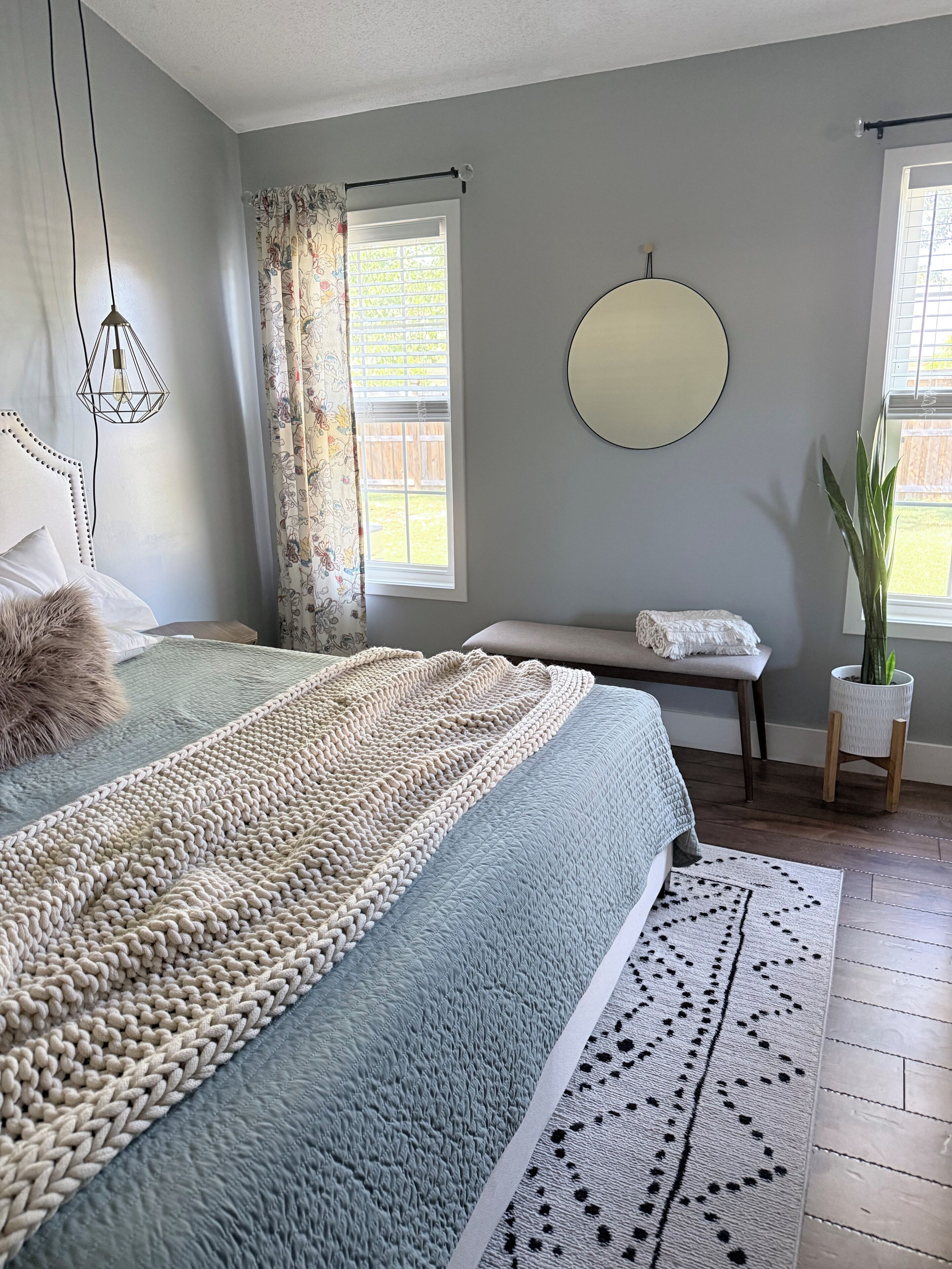 Une chambre à coucher avec un lit, une table de chevet, une plante en pot, un miroir rond et une fenêtre avec des rideaux.