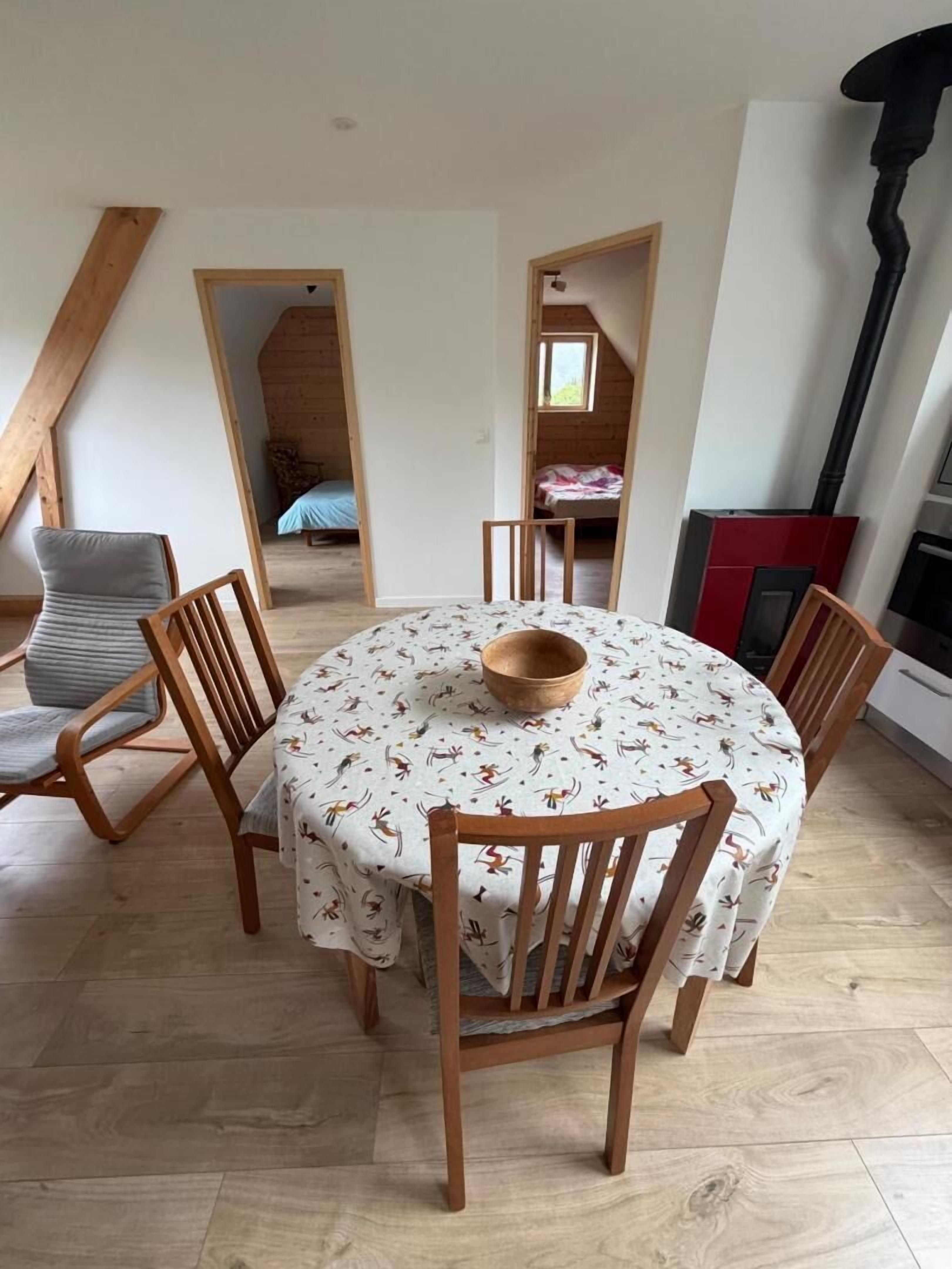 A dining area with a round table, wooden chairs, and a wooden bowl. There is a lofted sleeping area with a bed and a window.