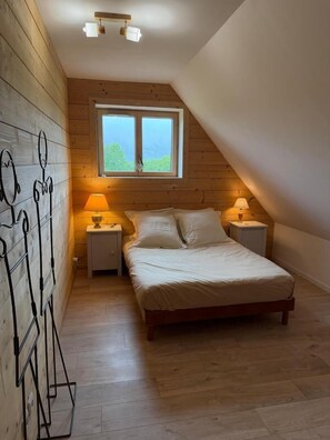 Ein Schlafzimmer mit Holzbalkendecke, einem Bett mit weißer Bettwäsche, zwei Nachttischlampen und einem Fenster mit Blick auf Grünflächen.