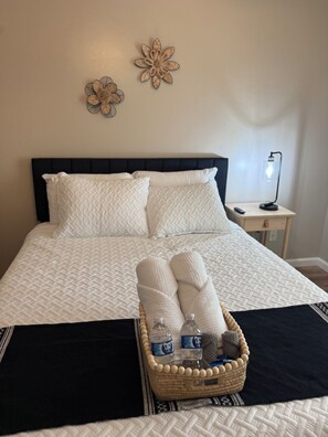 A neatly made bed with a black headboard, white pillows, and a quilted white bedspread. There is a small bedside table with a lamp and a decorative wall piece. A wicker basket with towels and bottles is placed on the bed.