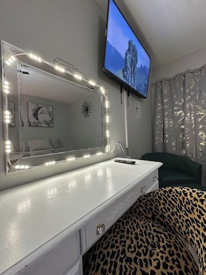 Quarto com espelho de toucador, televisão fixada na parede, almofada com padrão de leopardo e uma cadeira verde.