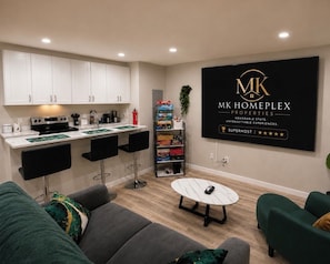 Una cocina moderna con armarios blancos, una encimera negra y una cafetera. Hay una zona de comedor con dos sillas negras y una mesa blanca. Un sofá verde y un sillón verde se encuentran en la sala de estar. Se observa una pared negra con el logo 'MK HOMEPLEX PROPERTIES' y una calificación de cinco estrellas.
