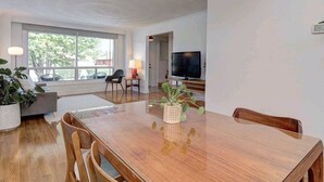 Un salon moderne avec une table à manger en bois, des chaises, un téléviseur et une grande fenêtre.