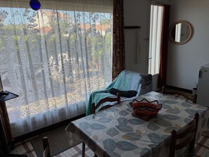 Une pi èce comprenant un lit, une table recouverte d’une nappe à motifs, une chaise, une fenêtre ornée de rideaux en dentelle, un miroir et un petit réfrigérateur.