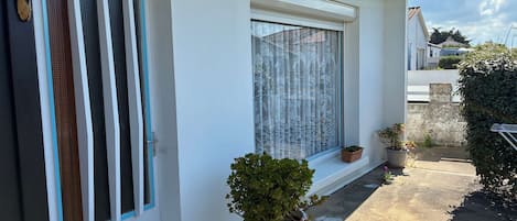 A white building with a blue roof, a window with lace curtains, and potted plants on the ground.