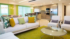 A modern living room with a sofa, armchairs, a glass coffee table, and a kitchen area with appliances.