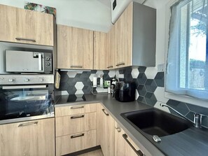 A modern kitchen with wooden cabinets, a microwave, an oven, a coffee maker, and a window with a view. - CAP D'AGDE - Plage Richelieu / Réf 418 (CA P D'AGDE)