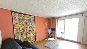 A living room with a sofa, a patterned tapestry, a fan, and a view through a glass door. - CAP D'AGDE - Plage du Môle / Réf 230 (CAP D'AGDE)