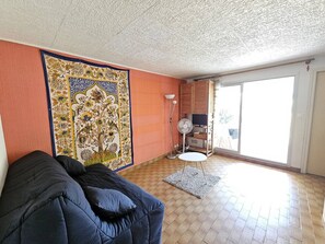 A living room with a sofa, a patterned tapestry, a fan, and a view through a glass door. - CAP D'AGDE - Plage du Môle / Réf 230 (CAP D'AGDE)