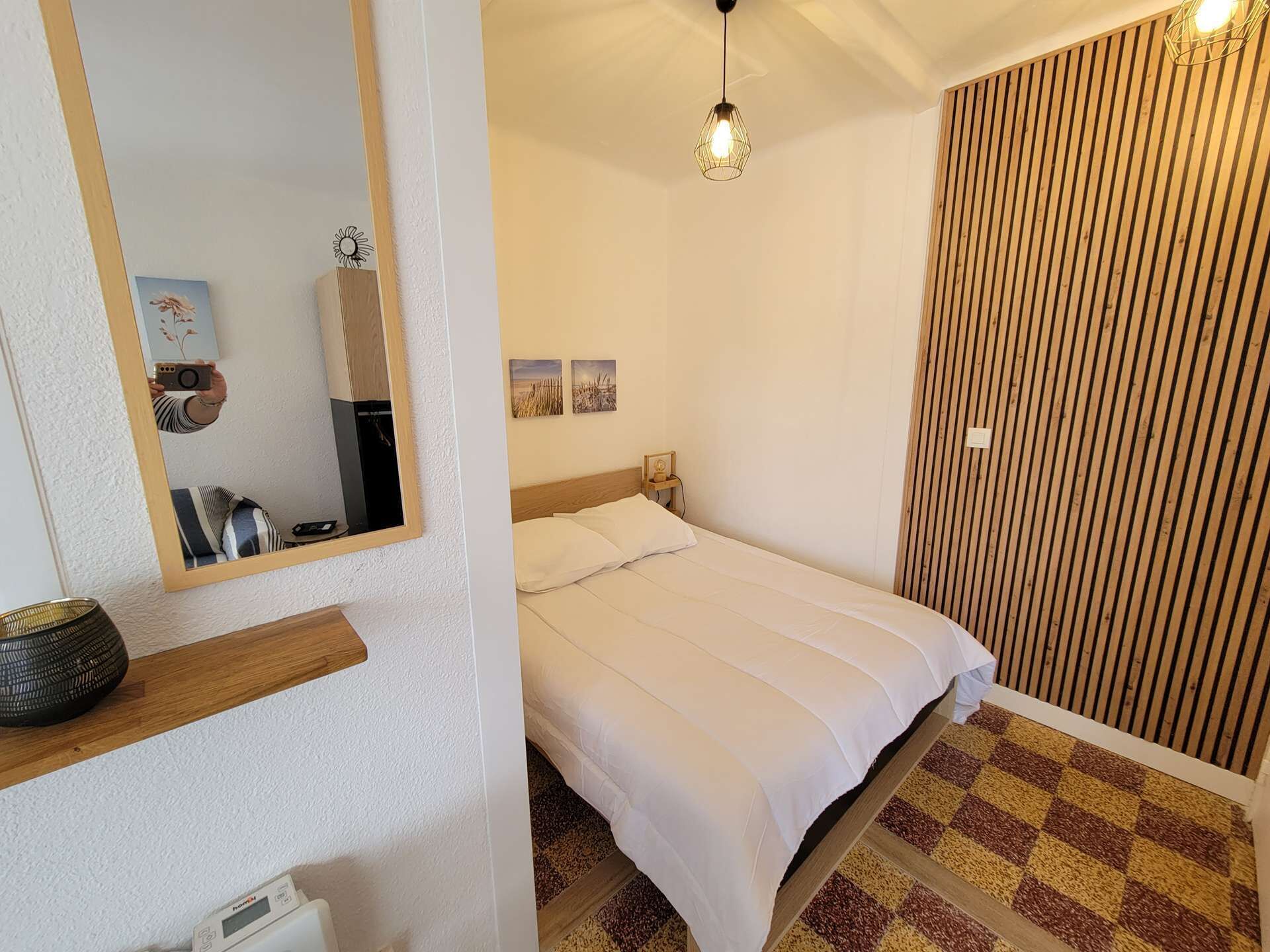 A bedroom with a bed, a mirror, a shelf with a decorative item, a wall-mounted light fixture, and a textured wall panel.