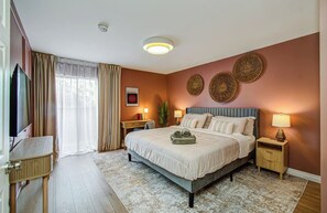 Una habitación de hotel con una cama, mesitas de noche, un televisor y un ventanal con cortinas.