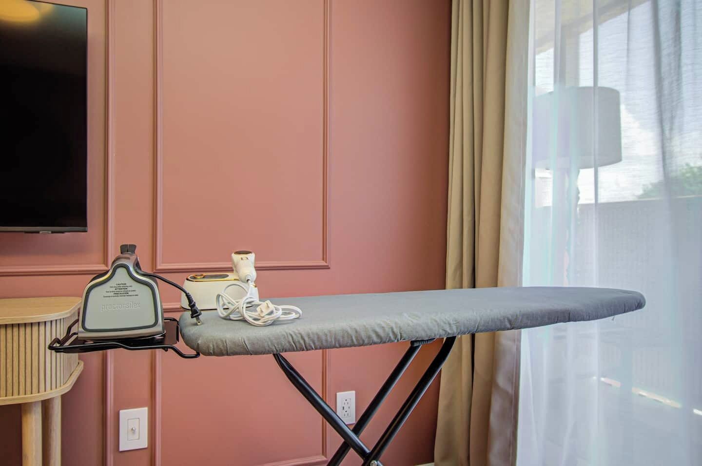 An ironing board with an iron and a power adapter in a room with pink walls and a window with curtains.