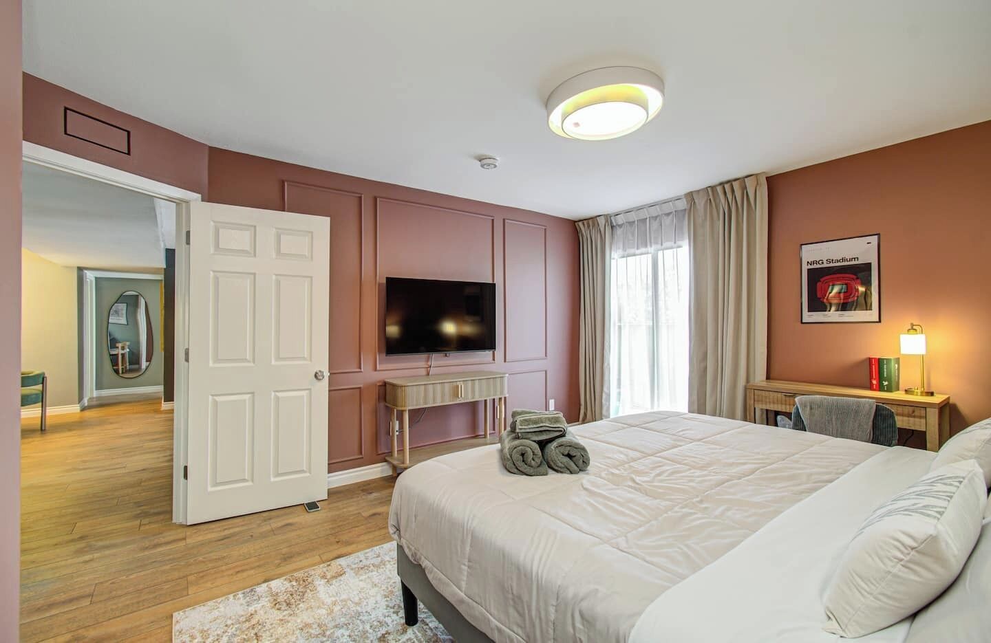 A hotel room with a large bed, a television, a desk with a lamp, and a view of a hallway.