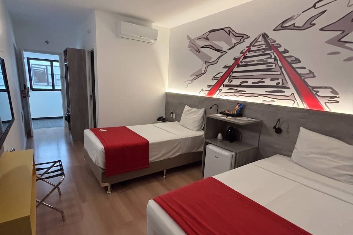 A hotel room with two beds, a desk, a chair, a small refrigerator, and a wall mural.