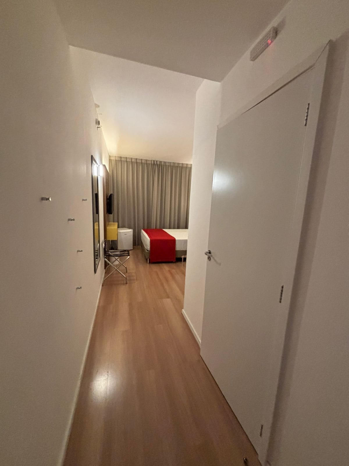 A narrow hallway leading to a bedroom with a bed, a red accent, and a mirror.