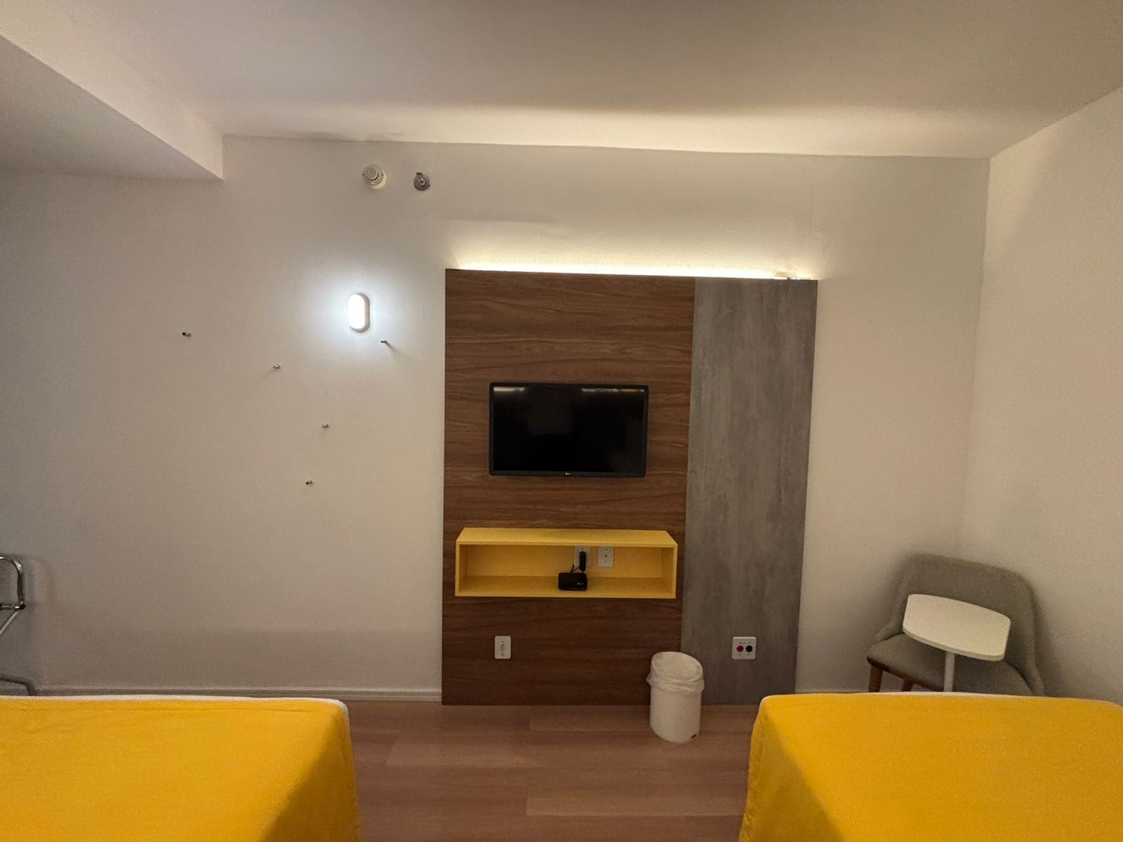 A hotel room with a TV mounted on a wooden panel wall, a small yellow shelf below it, a white trash bin, and two yellow beds.