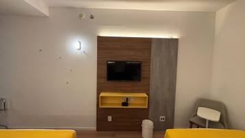 Habitación de hotel con un televisor montado en una pared de paneles de madera, un estante pequeño amarillo debajo, un cesto de basura blanco y dos camas amarillas.