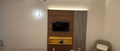 A hotel room with a TV mounted on a wooden panel wall, a small yellow shelf below it, a white trash bin, and two yellow beds.