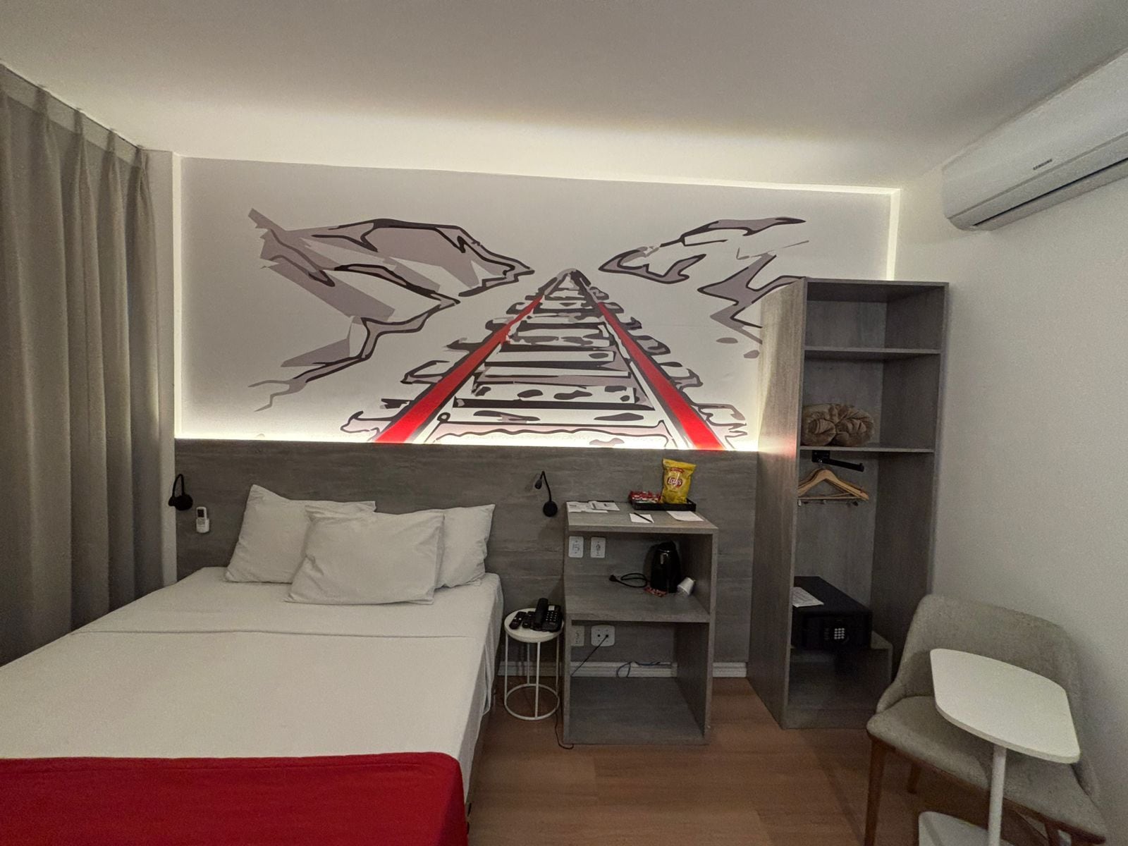 Una camera d'albergo con un letto, una scrivania con una sedia, un appendiabiti e un muro decorato con un murale raffigurante binari ferroviari.