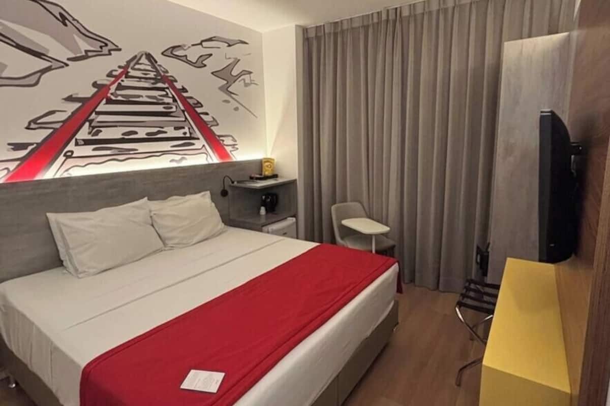 A hotel room with a bed, a desk, a chair, a TV, and a window with curtains.
