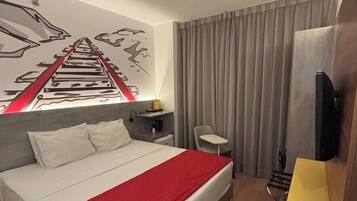 A hotel room with a bed, a desk, a chair, a TV, and a window with curtains.