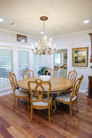 Une salle à manger avec une table ronde en bois, six chaises, un lustre et une vue sur l’extérieur grâce à de grandes fenêtres équipées de stores.