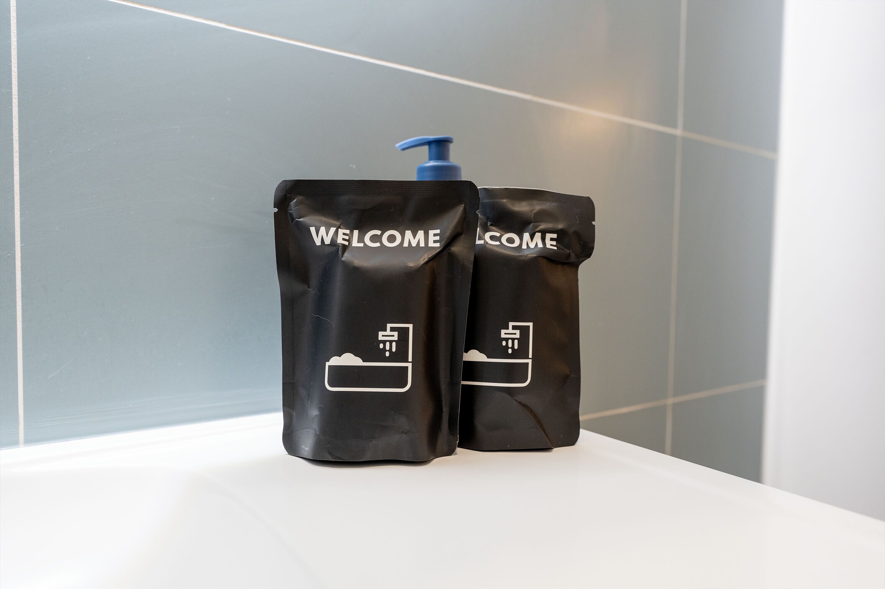 Two black welcome bags with a pump dispenser on a white surface against a tiled wall.