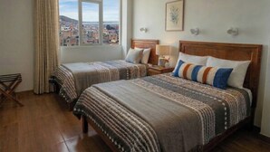 A hotel room with two beds, a window with a city view, a wooden floor, and a small table with a lamp.