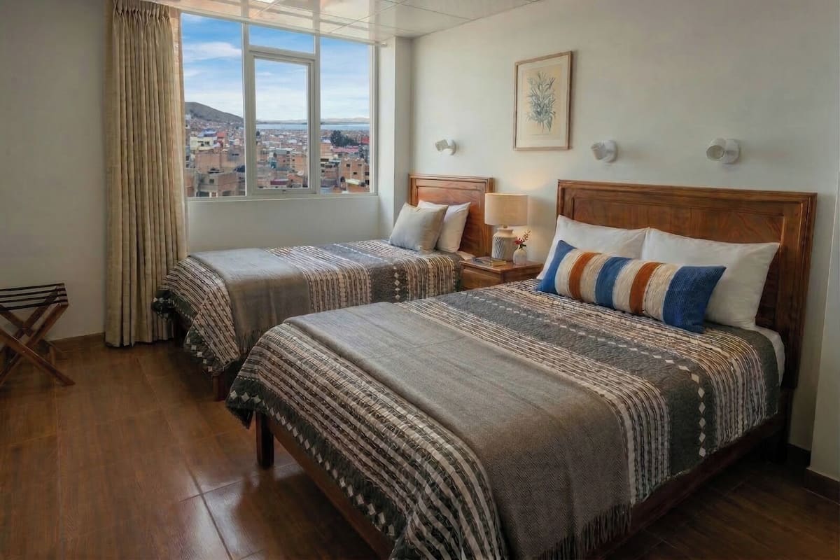 Habitación de hotel con dos camas, una ventana con vistas a la ciudad, piso de madera y una mesita con lámpara.