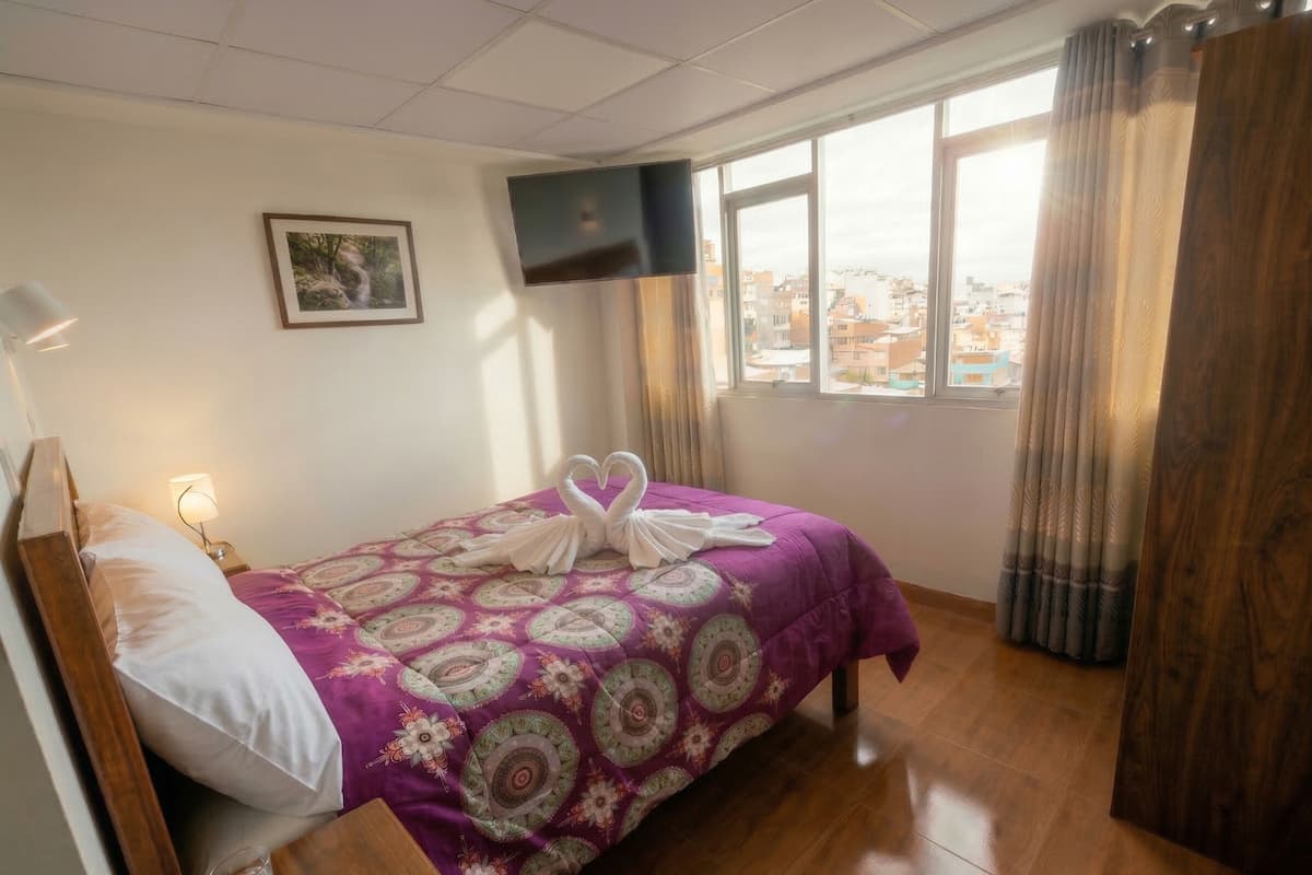 Habitación de hotel con cama, televisor en la pared, ventana con cortinas y vista a la ciudad.