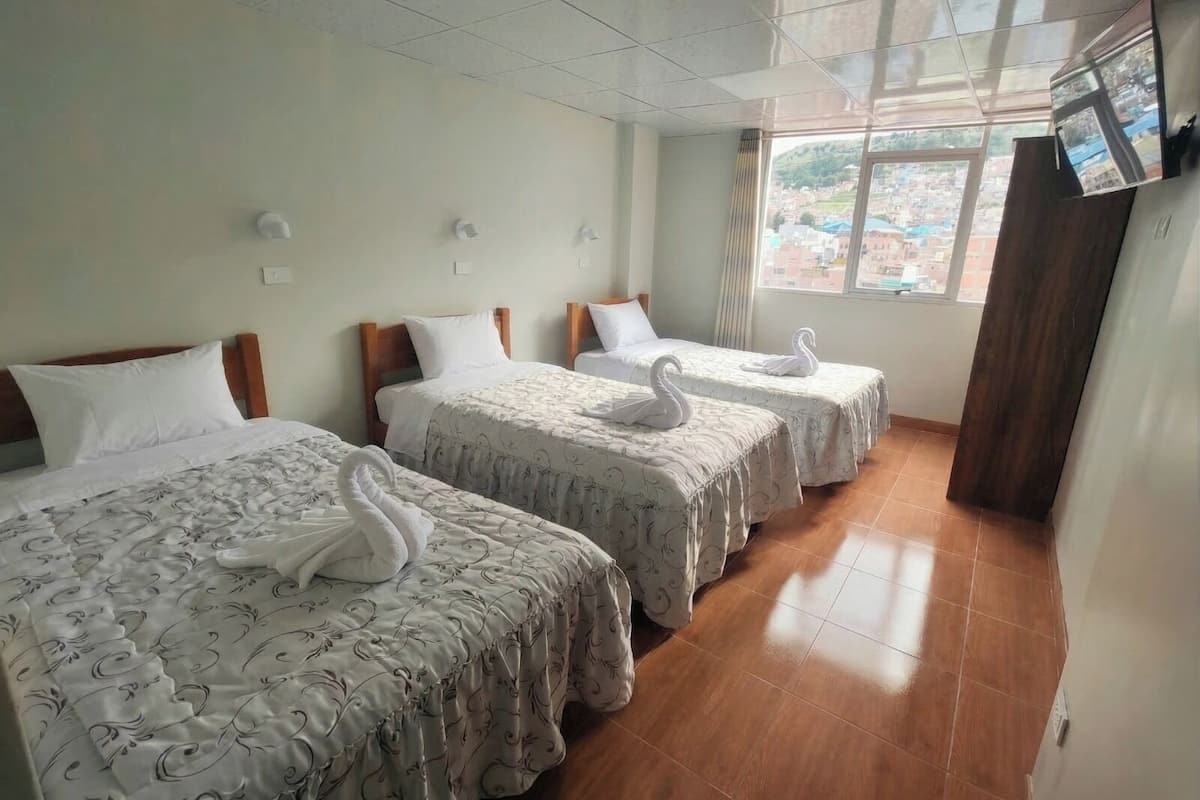 Habitación de hotel con tres camas, cada una con toalla con forma de cisne, una ventana con vistas a edificios y un televisor montado en la pared.