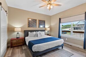A bedroom with a bed, nightstands, a ceiling fan, artwork, and a large window with a view. - Shore Thing | Steps to Pool | Hot Tub + Pool Table (Hollister)