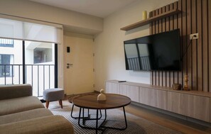 Ein modernes Wohnzimmer mit einer Couch, einem runden Tisch, einem an einer Holzwand montierten Fernseher und einer Schiebetür, die auf einen Balkon führt.