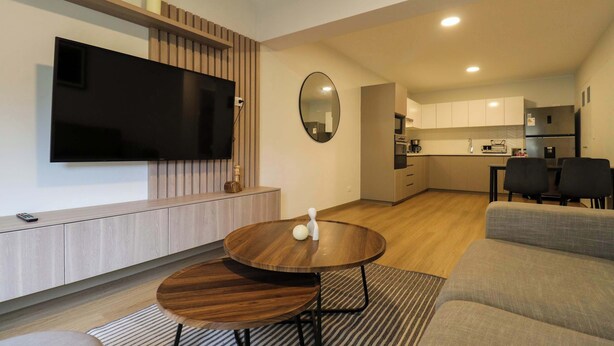Ein modernes Wohnzimmer mit Fernseher, Esstisch aus Holz, einer Couch und einer Küche im Hintergrund.
