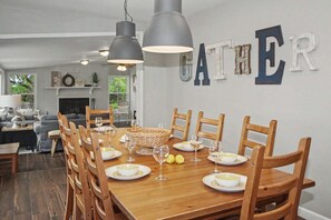 Ein Essbereich mit einem Holztisch, gedeckt für sechs Personen, umgeben von Holzstühlen, mit dem an der Wand befestigten Wort 'GATHER'.