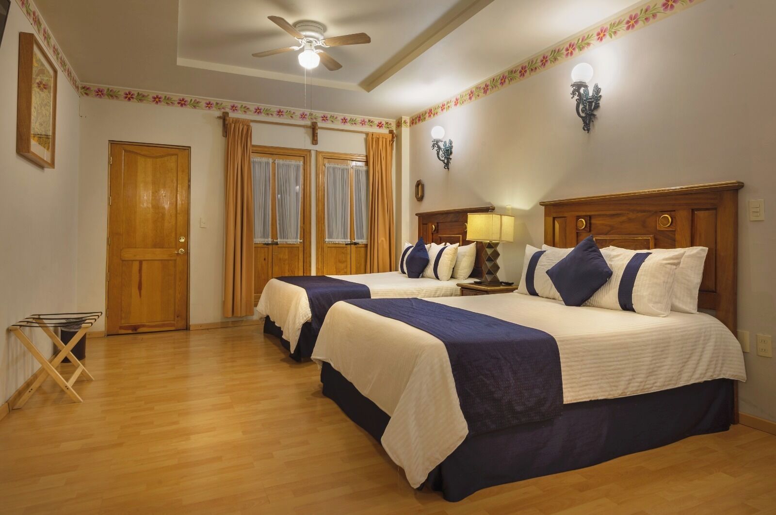 Habitación de hotel con dos camas, suelo de madera, ventilador de techo y una mesita con lámpara.