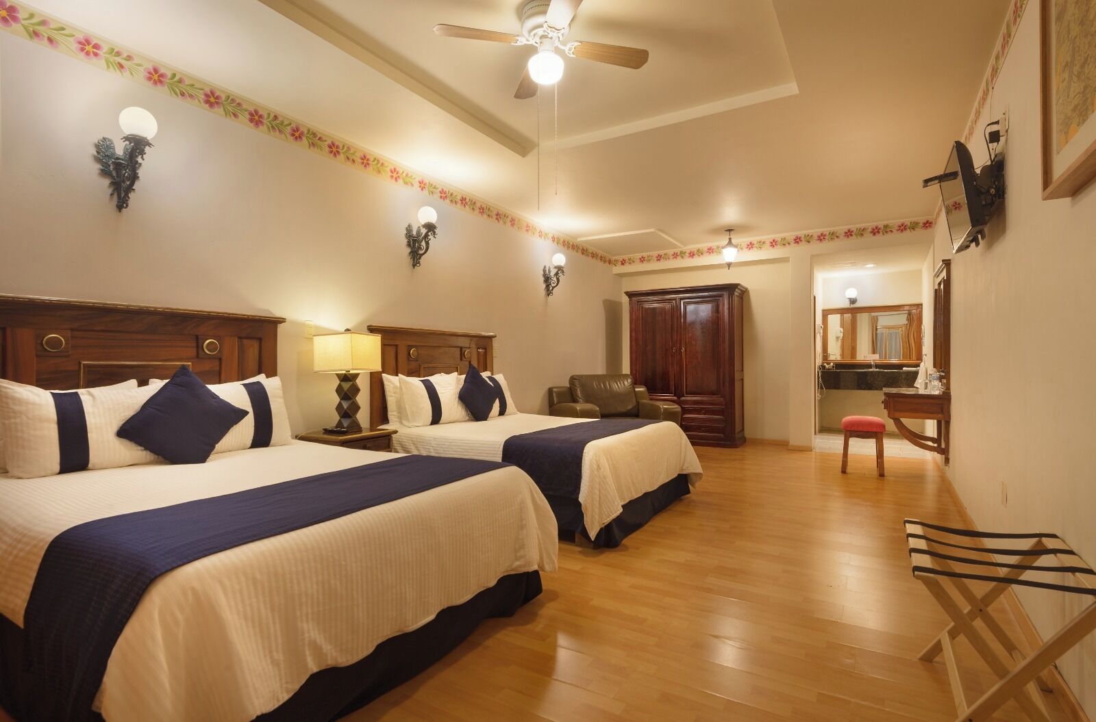 A hotel room with two beds, a ceiling fan, a desk with a chair, a mirror, and a wardrobe.