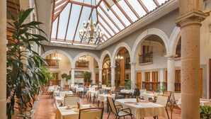 A spacious indoor dining area with high ceilings, arches, and a glass roof. Tables are set with chairs, and potted plants are placed around the room. - HOTEL BOUTIQUE GRAND MAYES (Pátzcuaro)