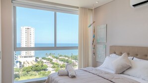Un dormitorio con un ventanal grande, una cama con ropa de cama blanca y vistas a la ciudad y al océano.