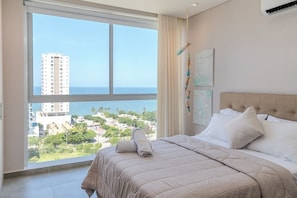 A bedroom with a large window, a bed with white bedding, and a view of a cityscape and ocean. - Salguero Suites by RV Stays (Santa Marta)