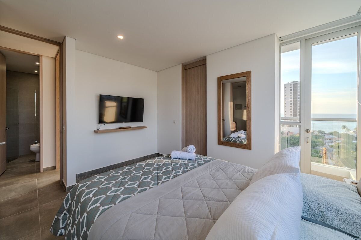 A modern hotel room with a large bed, a TV mounted on the wall, a balcony with a city view, and a sliding door leading to a bathroom.