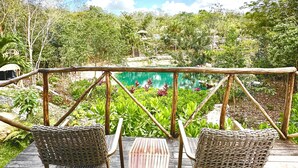Una terraza de madera con sillas de mimbre y una mesa, con vista a una piscina y abundante vegetación.