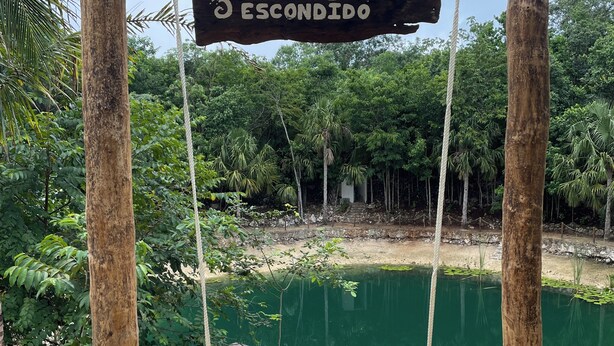 Un columpio de madera con un cartel que dice 'CENOTE ESCONDIDO' en un entorno natural con agua y follaje.