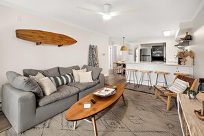 A modern living room with a grey sofa, wooden coffee table, and a surfboard on the wall. - Beautiful 3 Bedroom at Sea Breeze! (Ocean City)
