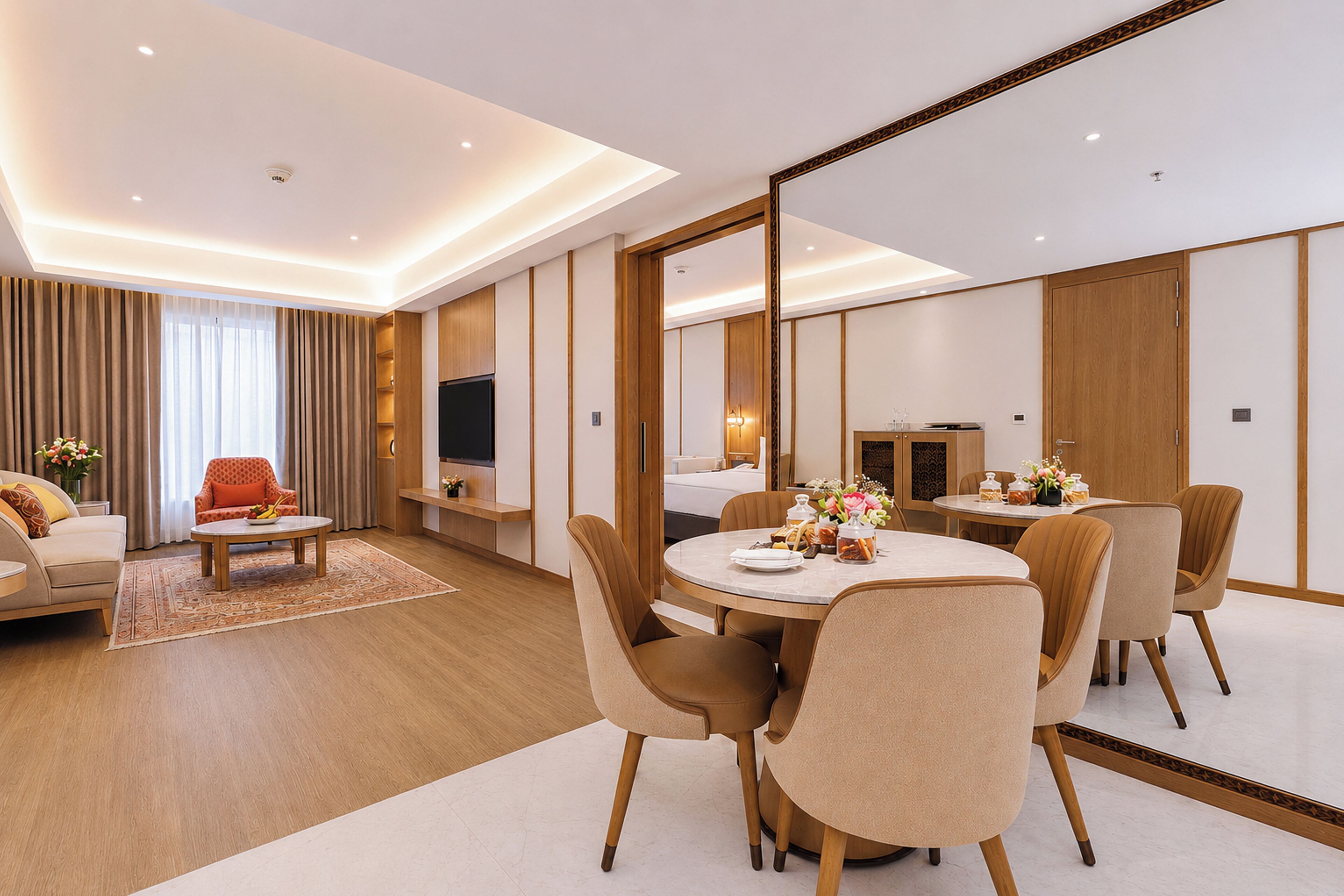 A modern hotel room with a dining area, a sofa, a TV, and a large mirror.
