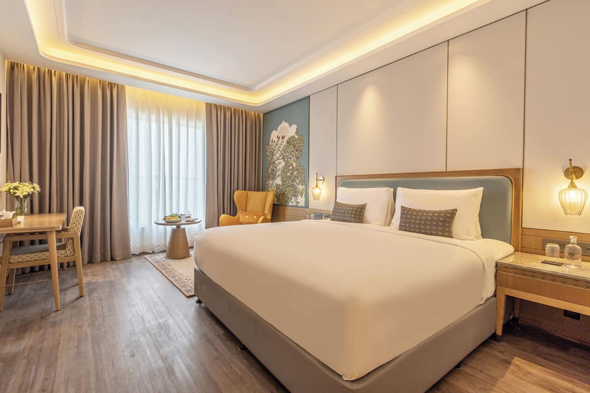 Una habitación de hotel con una cama grande, una mesita de noche con lámpara, una silla, una pequeña mesa con un florero y una ventana con cortinas.