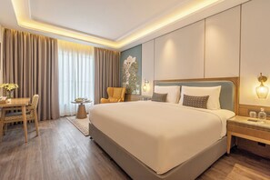 Una habitación de hotel con una cama grande, una mesita de noche con lámpara, una silla, una pequeña mesa con un florero y una ventana con cortinas.